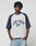 A man with braided hair wears the Live Fast Raglan Tee by LIVE FAST DIE YOUNG—an oversized white and dark blue raglan t-shirt. He stands with hands in his pockets, looking at the camera with a neutral expression.