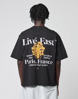 A person with short, twisted hair faces away, wearing the LIVE FAST DIE YOUNG LFDY Paris Tee in Oversize Fit. The black shirt features bold white text, a yellow Paris graphic, French phrases, a floral design, and prominent front and back prints.