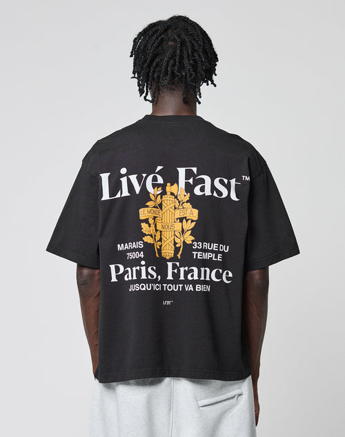 A person with short, twisted hair faces away, wearing the LIVE FAST DIE YOUNG LFDY Paris Tee in Oversize Fit. The black shirt features bold white text, a yellow Paris graphic, French phrases, a floral design, and prominent front and back prints.