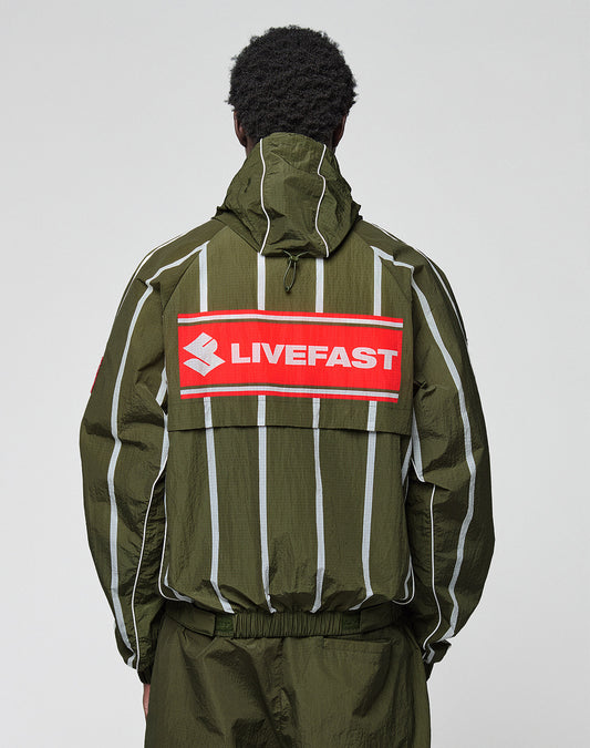A person seen from behind wears the LFDY Nylon Trackjacket by LIVE FAST DIE YOUNG—an olive green, oversize jacket with white vertical stripes, a red patch on the back reading "LIVEFAST," and a hood with elastic drawstring.