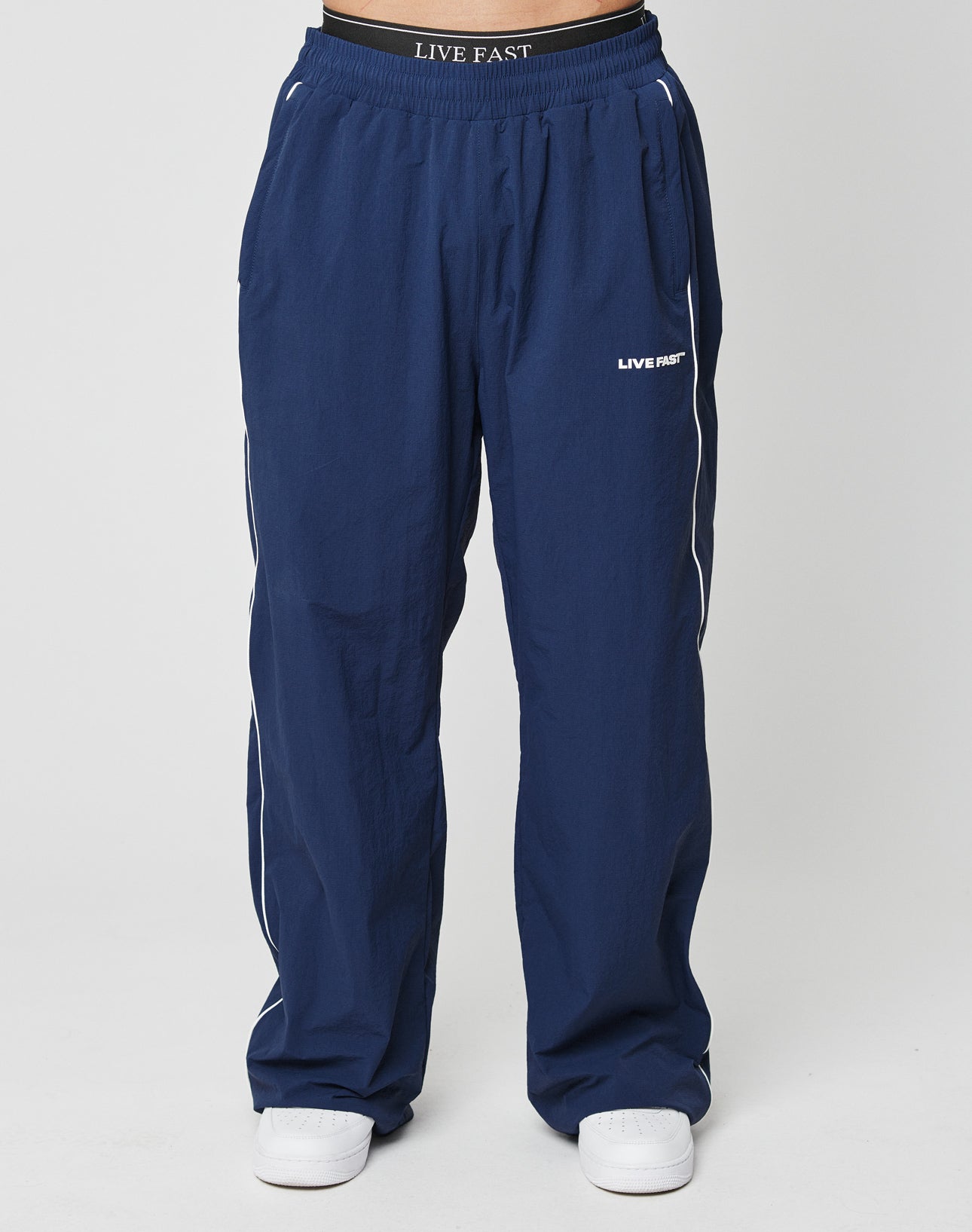 A person wearing LIVE FAST DIE YOUNG LFDY Ripstop Trackpants in navy blue with white side stripes, silicone LIVE FAST print on the left thigh, wide fit and elastic waistband, paired with white shoes against a white background.