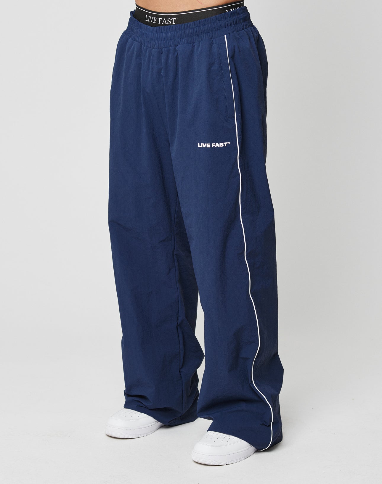 Model wears wide fit navy LFDY Ripstop Trackpants by LIVE FAST DIE YOUNG, featuring an elastic waistband, white side piping, and LIVE FAST silicone print on the waistband and left thigh, styled with white sneakers against a light background.