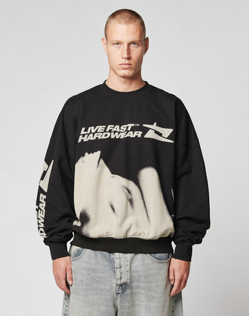 A man with short hair wears the LIVE FAST DIE YOUNG LFDY Mystic Sweater—a black, oversized cotton sweatshirt with a large face graphic and bold text—paired with faded blue jeans. He stands against a plain, light background.