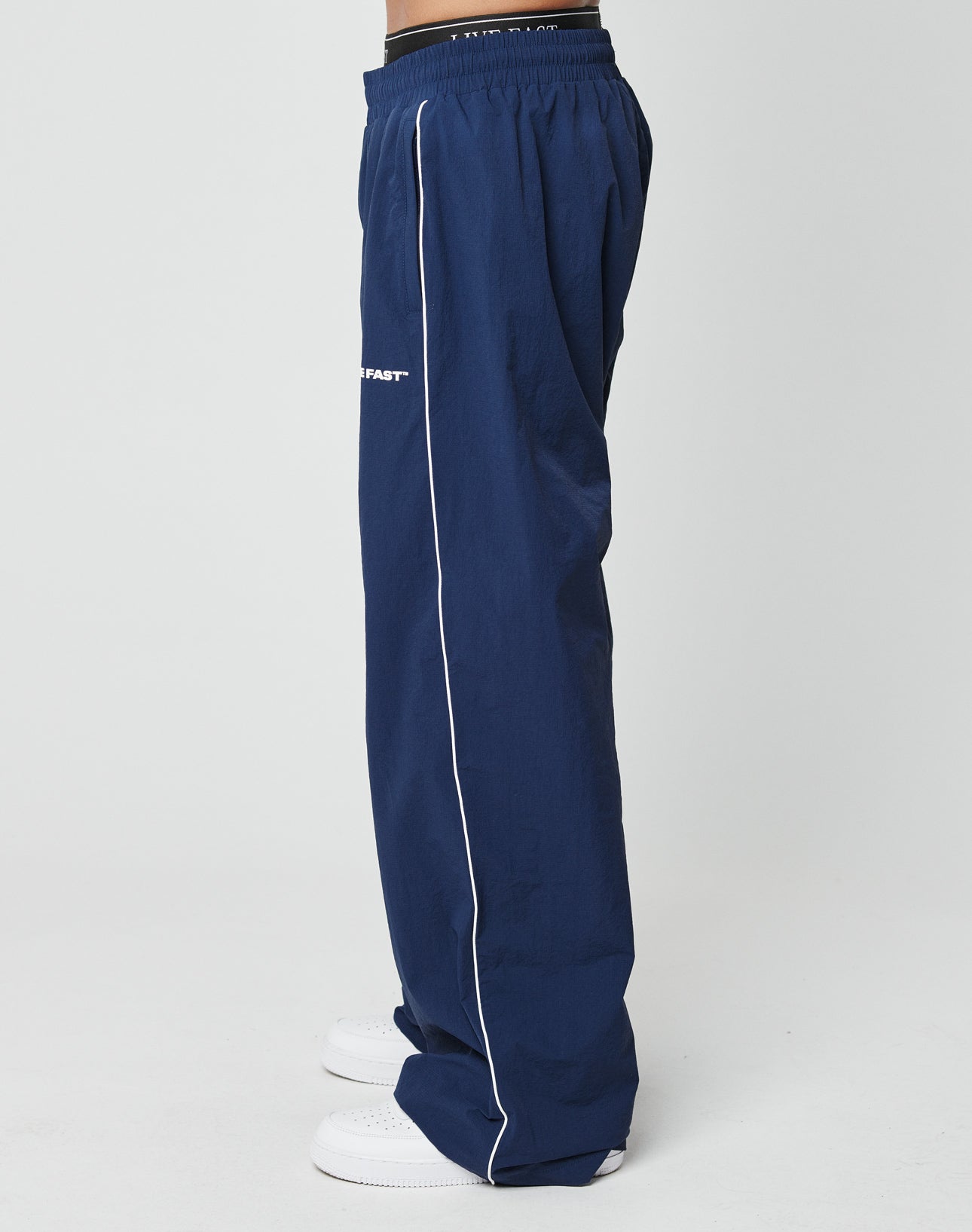 A person stands sideways against a white background wearing LIVE FAST DIE YOUNG LFDY Ripstop Trackpants in wide fit navy blue with white piping and an elastic waistband, paired with white sneakers.
