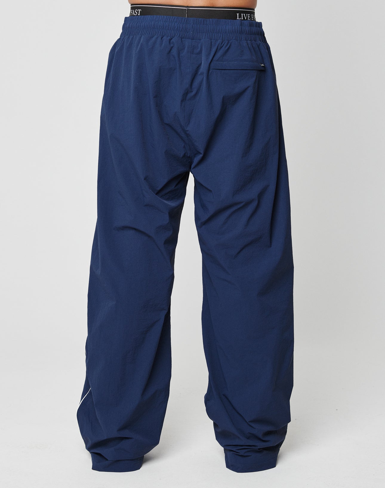 A person is seen from behind wearing LIVE FAST DIE YOUNG LFDY Ripstop Trackpants in loose-fit navy blue, featuring an elastic waistband and a right-side back pocket. The background is plain and light-colored.