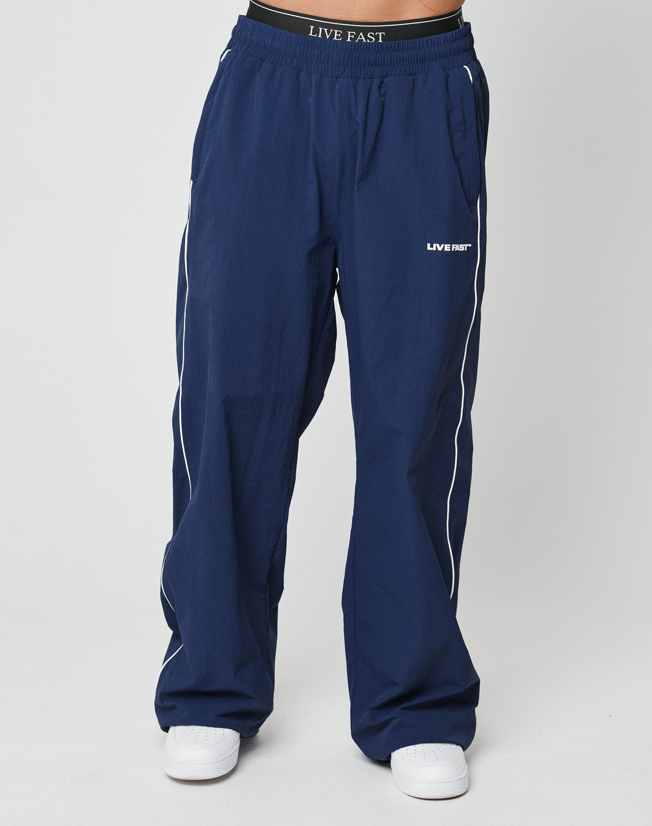 A person wearing LFDY Ripstop Trackpants by LIVE FAST DIE YOUNG, featuring navy blue color, white piping, an elastik-bund waistband with Silikonprint LIVE FAST, and white sneakers stands against a plain light background.