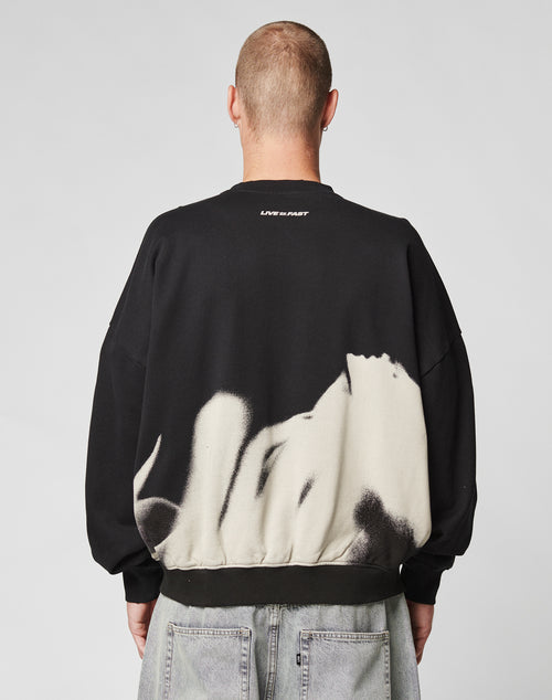 A person with a shaved head faces away wearing the LIVE FAST DIE YOUNG LFDY Mystic Sweater, an oversized black cotton sweatshirt with a white abstract back pattern, topstitching, small white neckline text, and paired with light gray jeans.
