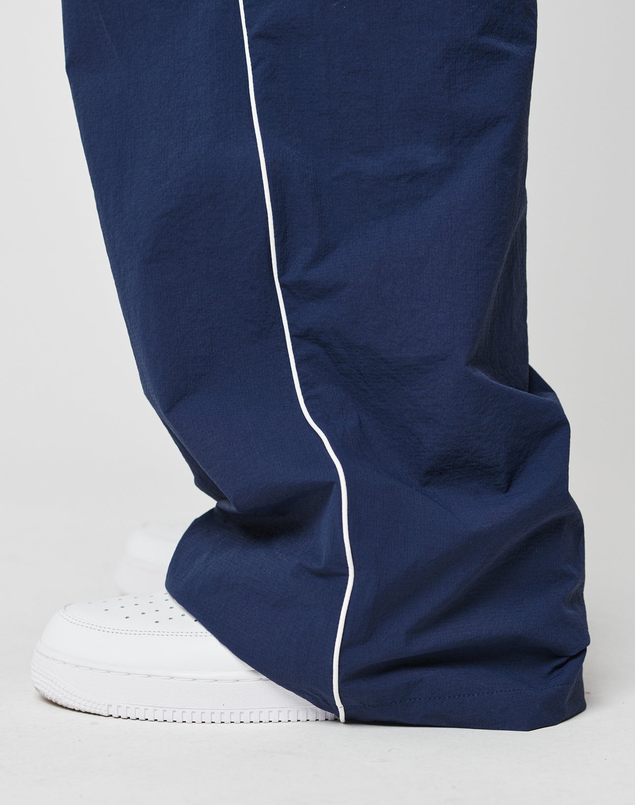 A close-up of a person wearing LFDY Ripstop Trackpants by LIVE FAST DIE YOUNG in navy with white piping, featuring an elastic waistband and wide fit, paired with white sneakers on a light-colored surface.