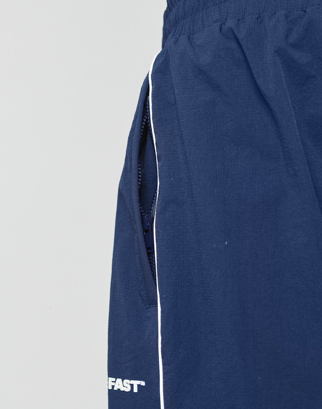Close-up of LIVE FAST DIE YOUNG LFDY Ripstop Trackpants in navy with a white side stripe, elastic waistband, and partial zipper pocket. “FAST” is featured near the hem in silicone print.