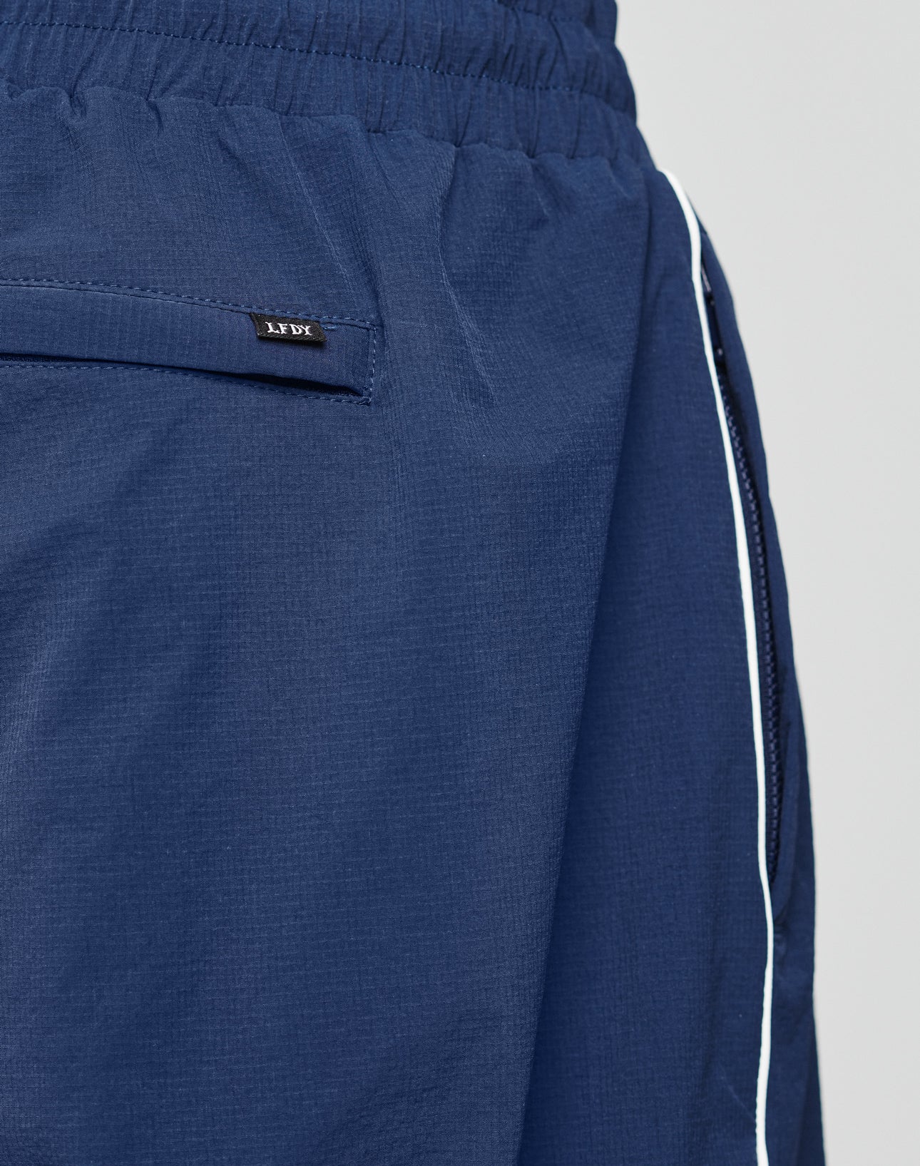 Close-up of LFDY Ripstop Trackpants by LIVE FAST DIE YOUNG in blue, featuring a zippered pocket, white side piping, comfortable Elastik-Bund, wide fit, and slightly textured ripstop fabric.