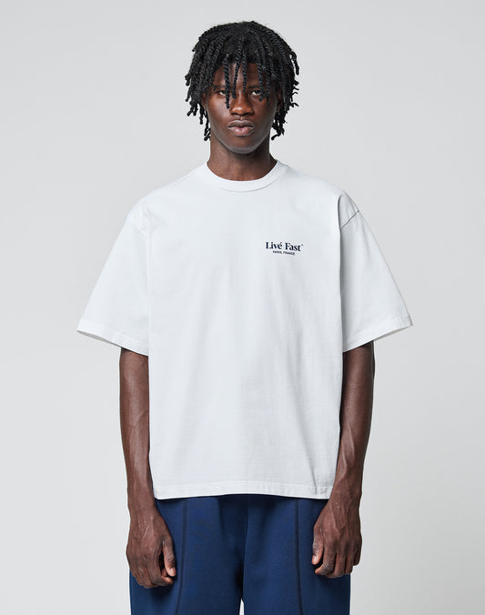 A person with medium-length dreadlocks wears the LFDY Paris Tee by LIVE FAST DIE YOUNG, an oversized white cotton shirt with front and back print, paired with dark blue pants, standing against a plain light gray background.