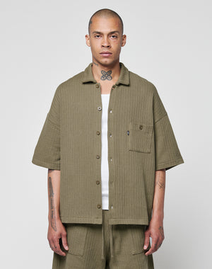 A person with a shaved head and visible tattoos stands facing forward, wearing the LIVE FAST DIE YOUNG Basic Knit Shirt in olive green with a Hemdkragen and Brusttasche, paired with wide-legged matching shorts against a plain light gray background.