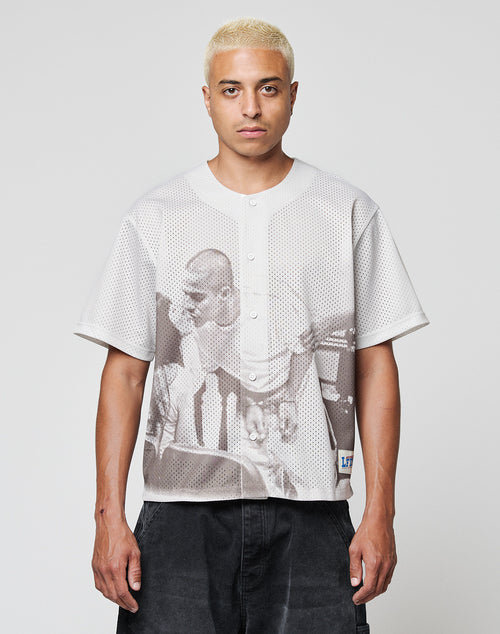 A person with short, bleached blond hair wears the LIVE FAST DIE YOUNG LFDY Courtroom Jersey, an oversized light short-sleeve shirt with a large grayscale print of two people, standing facing the camera against a plain background.