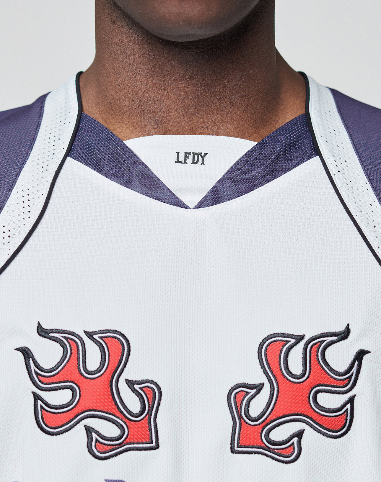 A person wearing the LIVE FAST DIE YOUNG LFDY Call Us Jersey—a white polyester sports top with blue trim, red flame chest designs, and a V-Ausschnitt. Only the lower face, neck, and upper torso are visible.