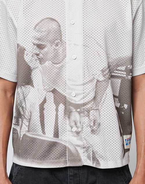 A close-up of a person wearing the LIVE FAST DIE YOUNG LFDY Courtroom Jersey, an oversize fit white mesh button-up shirt featuring an all-over grayscale print of two people and a rectangular white patch with blue text on the lower side.
