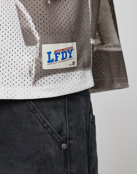 A close-up shows the lower part of the oversize LFDY Courtroom Jersey by LIVE FAST DIE YOUNG, featuring perforated patterned fabric and a sewn patch labeled LFDY OFFICIALLY LICENSED PRODUCT, paired with dark gray pants.