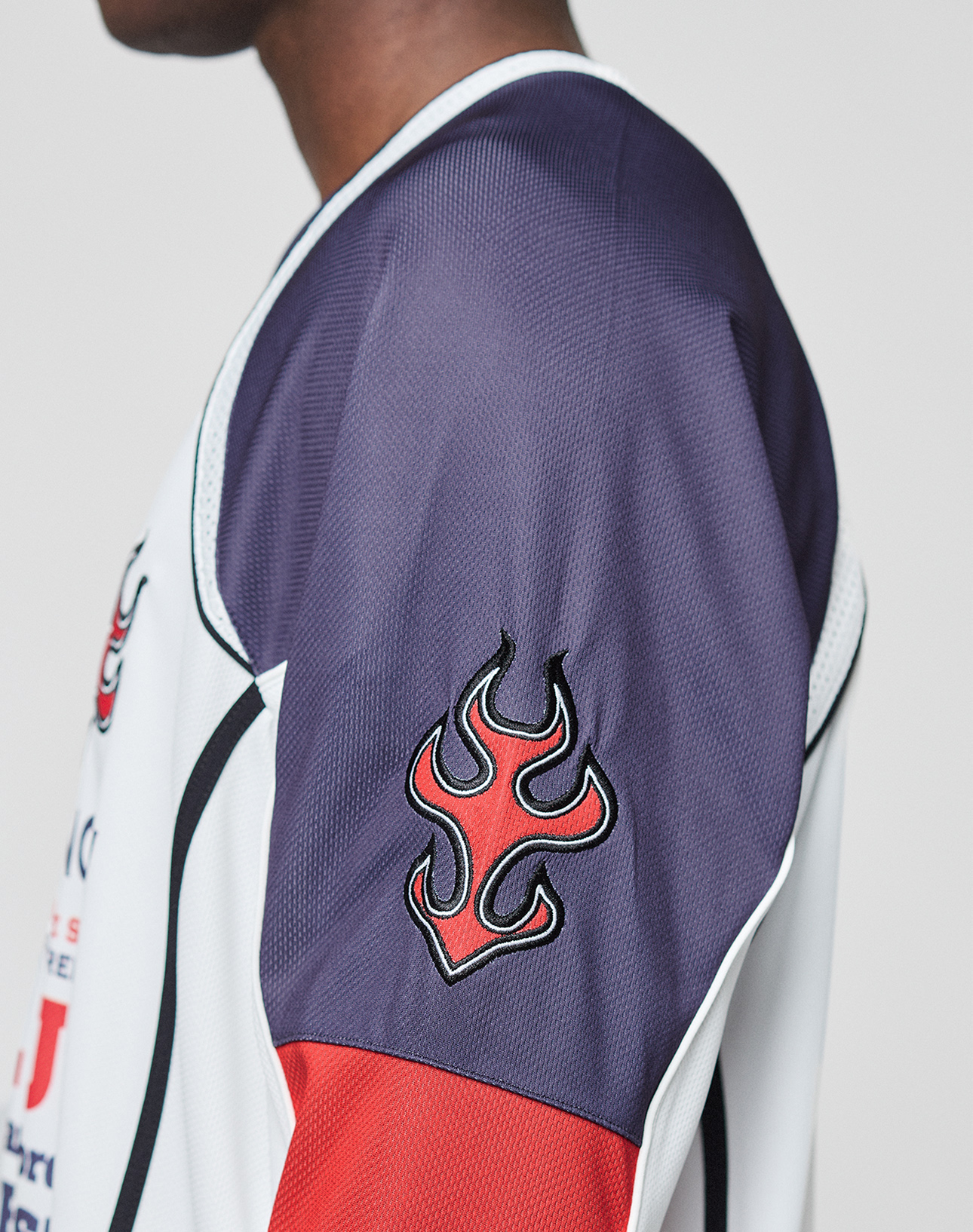 A close-up side view of someone wearing the LFDY Call Us Jersey by LIVE FAST DIE YOUNG, featuring dark purple, white, and red polyester panels, a bold flame logo on the shoulder, and a sporty V-Ausschnitt. The face is partially out of frame.
