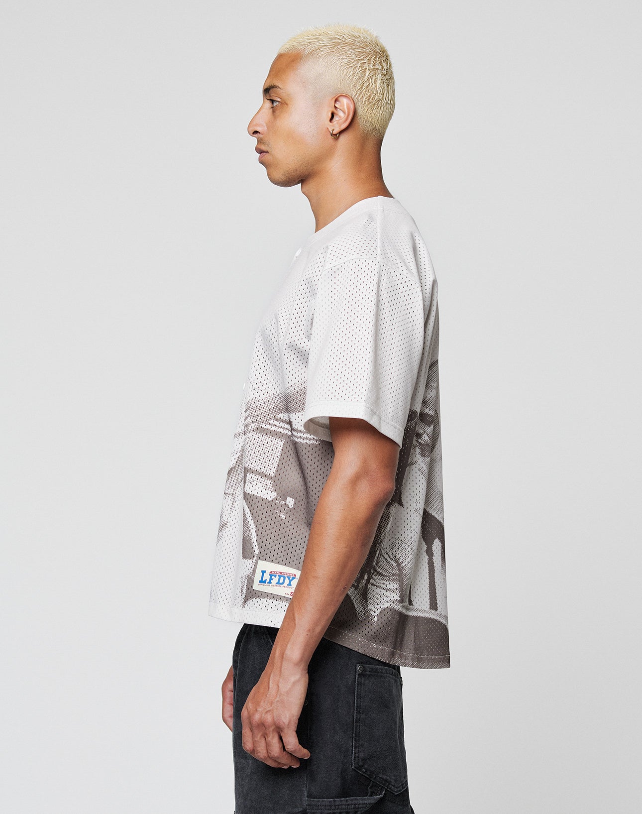 A person with short, bleached hair stands in profile, wearing the LFDY Courtroom Jersey by LIVE FAST DIE YOUNG—an oversize graphic tee with a small LFDY patch at the hem—paired with dark jeans against a plain background.
