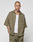 A person with a shaved head and tattoos wears the LIVE FAST DIE YOUNG Basic Knit Shirt in olive green, styled oversized over a white tank and matching pants, standing against a plain light gray background.