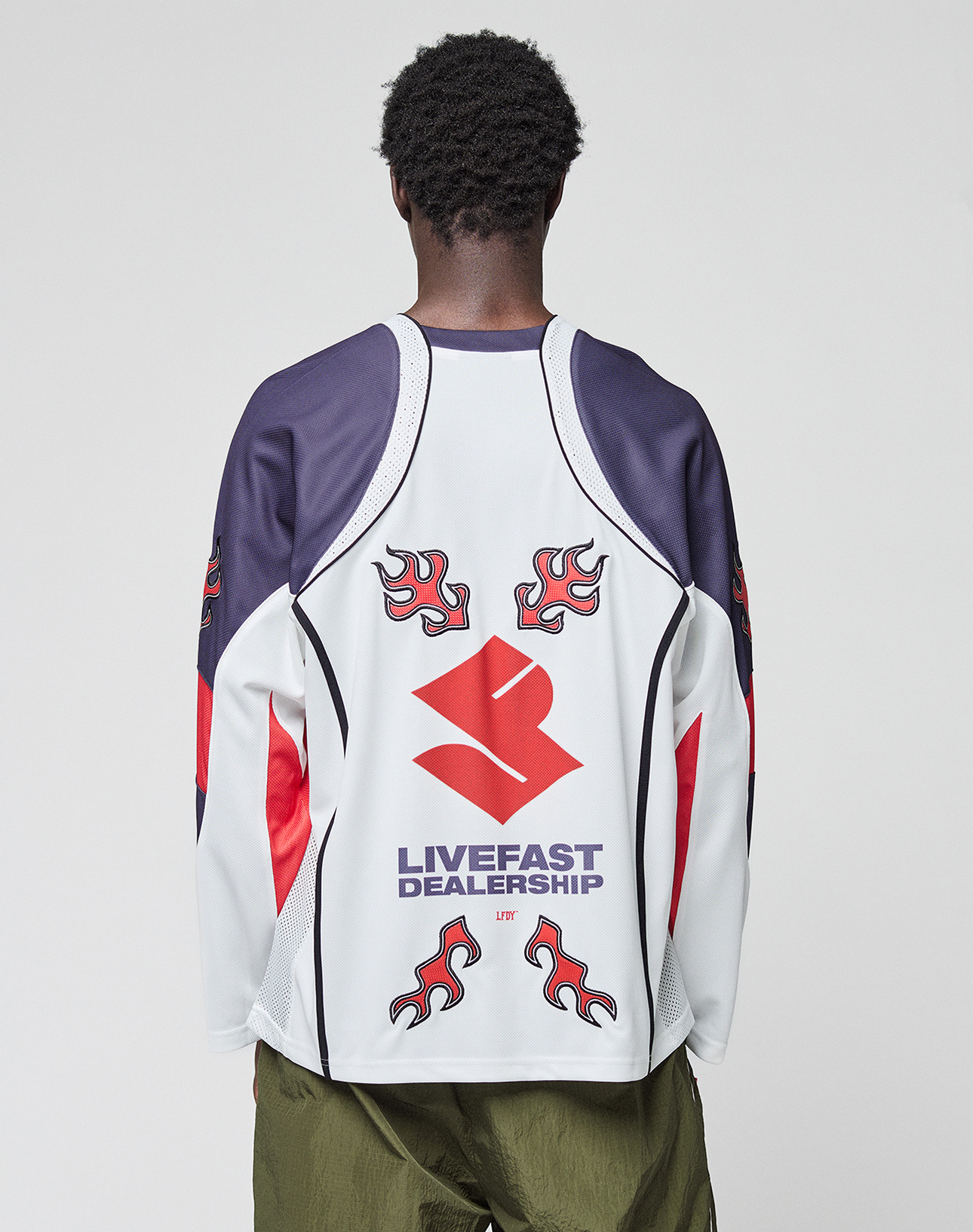 Seen from behind, a person with short curly hair wears the LFDY Call Us Jersey by LIVE FAST DIE YOUNG—an oversized white and navy long-sleeve top with red flame graphics, a bold red logo, and LIVEFAST DEALERSHIP text on the back.