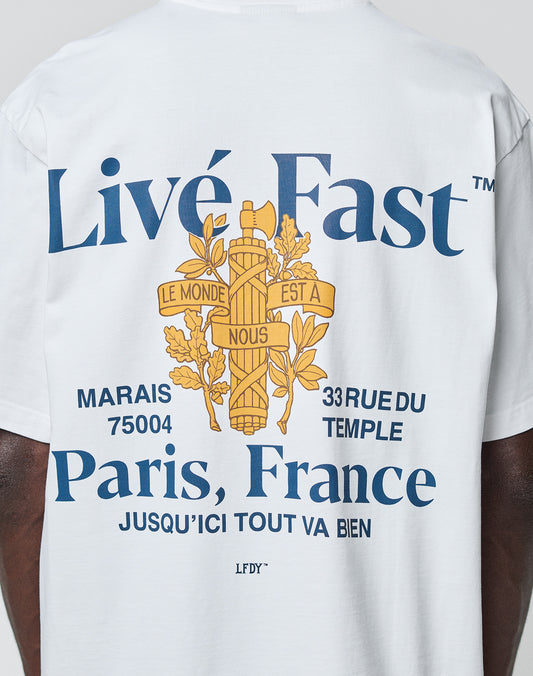 Someone wears the LFDY Paris Tee by LIVE FAST DIE YOUNG—an oversize white cotton shirt with blue Livé Fast and Paris, France text, ornate yellow cross graphics, French words, and 33 Rue du Temple, Marais 75004 on front and back.