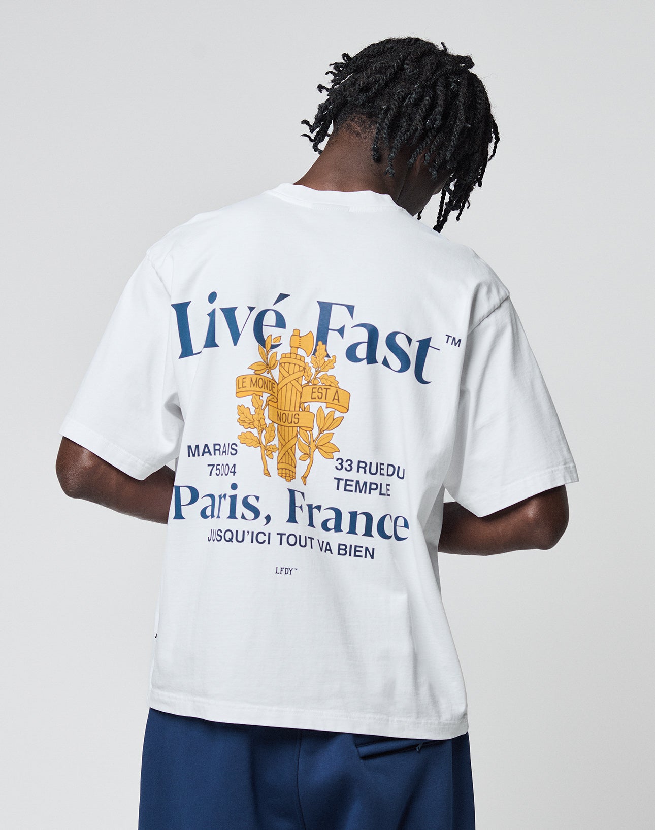 A person with short dreadlocks faces away, wearing the LIVE FAST DIE YOUNG LFDY Paris Tee—an Oversize Fit white T-shirt featuring bold blue Live Fast Paris, France text, a gold crest, and French phrases on front and back.