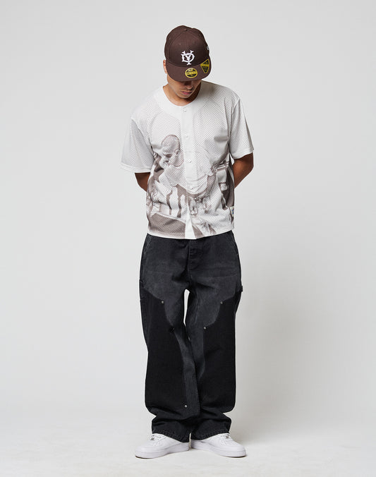 A person wearing a brown baseball cap, loose black jeans, white sneakers, and the LIVE FAST DIE YOUNG LFDY Courtroom Jersey stands against a plain white background, looking down with hands behind their back.