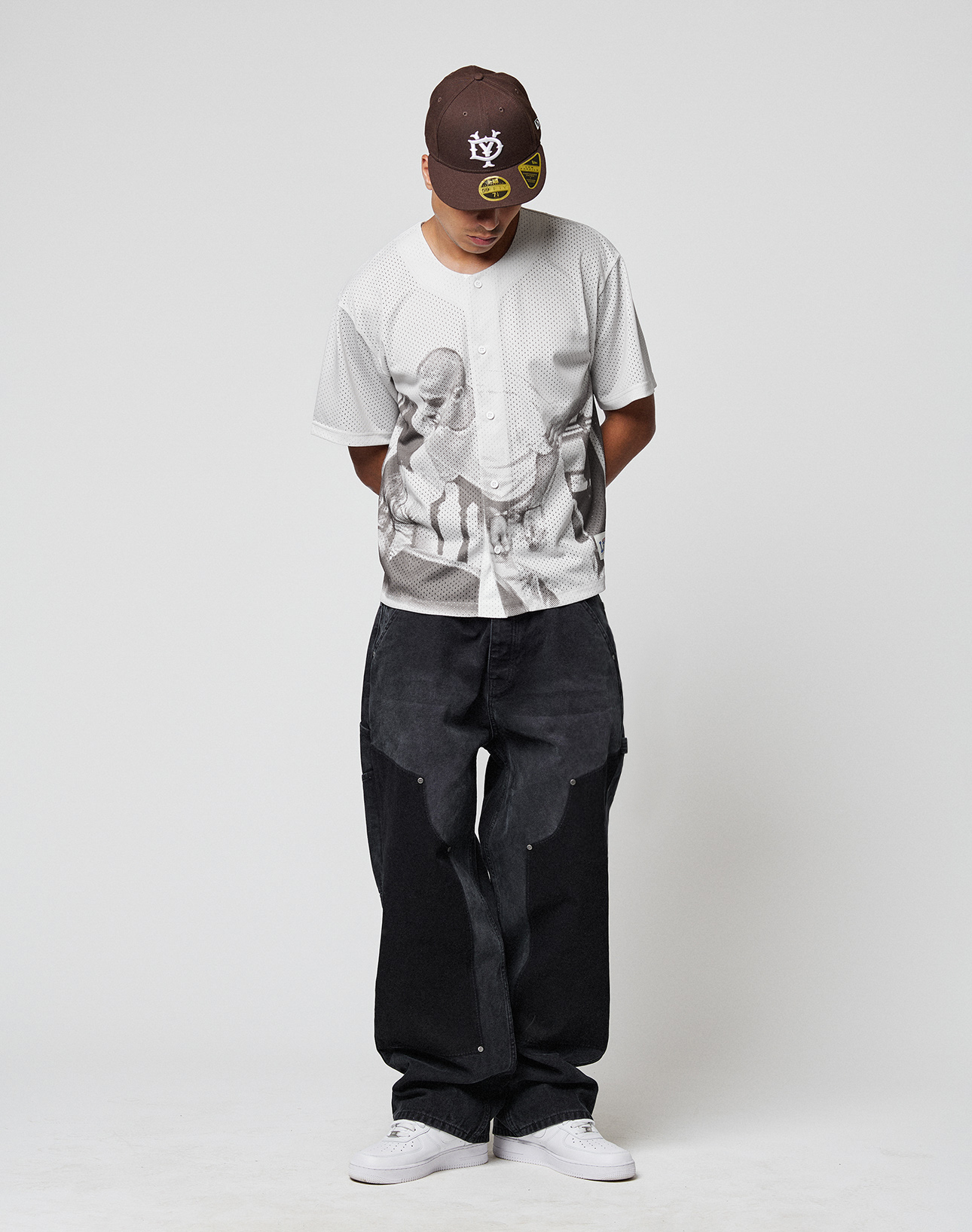 A man stands against a plain white background, looking down with his hands behind his back, wearing the LFDY Courtroom Jersey by LIVE FAST DIE YOUNG, loose black jeans, an oversize brown cap with yellow graphics, and white sneakers.