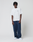 A man with short dreadlocks stands against a plain background, wearing a loose white logo T-shirt, LIVE FAST DIE YOUNG LFDY Active Fleece Jogger pants in navy blue, and white sneakers. He faces forward with arms relaxed by his sides.