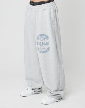 A person wears Loyal Wide Jogger sweatpants by LIVE FAST DIE YOUNG in light gray, featuring a wide leg, elastic waistband, and graphic on the upper thigh. Black underwear is visible above the pants, paired with white sneakers.