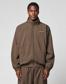 A person faces the camera against a plain gray background, wearing the brown polyester LFDY Trackjacket by LIVE FAST DIE YOUNG with an oversize fit and LIVE FAST text on the left chest, paired with matching pants. | model