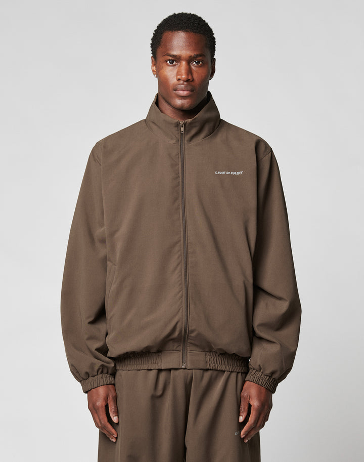 A person faces the camera against a plain gray background, wearing the brown polyester LFDY Trackjacket by LIVE FAST DIE YOUNG with an oversize fit and LIVE FAST text on the left chest, paired with matching pants. | model
