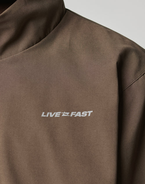 Close-up of the LFDY Trackjacket by LIVE FAST DIE YOUNG, a brown oversized polyester jacket with LIVE FAST in white capitals, a small stylized runner, and reflective print on the left chest.