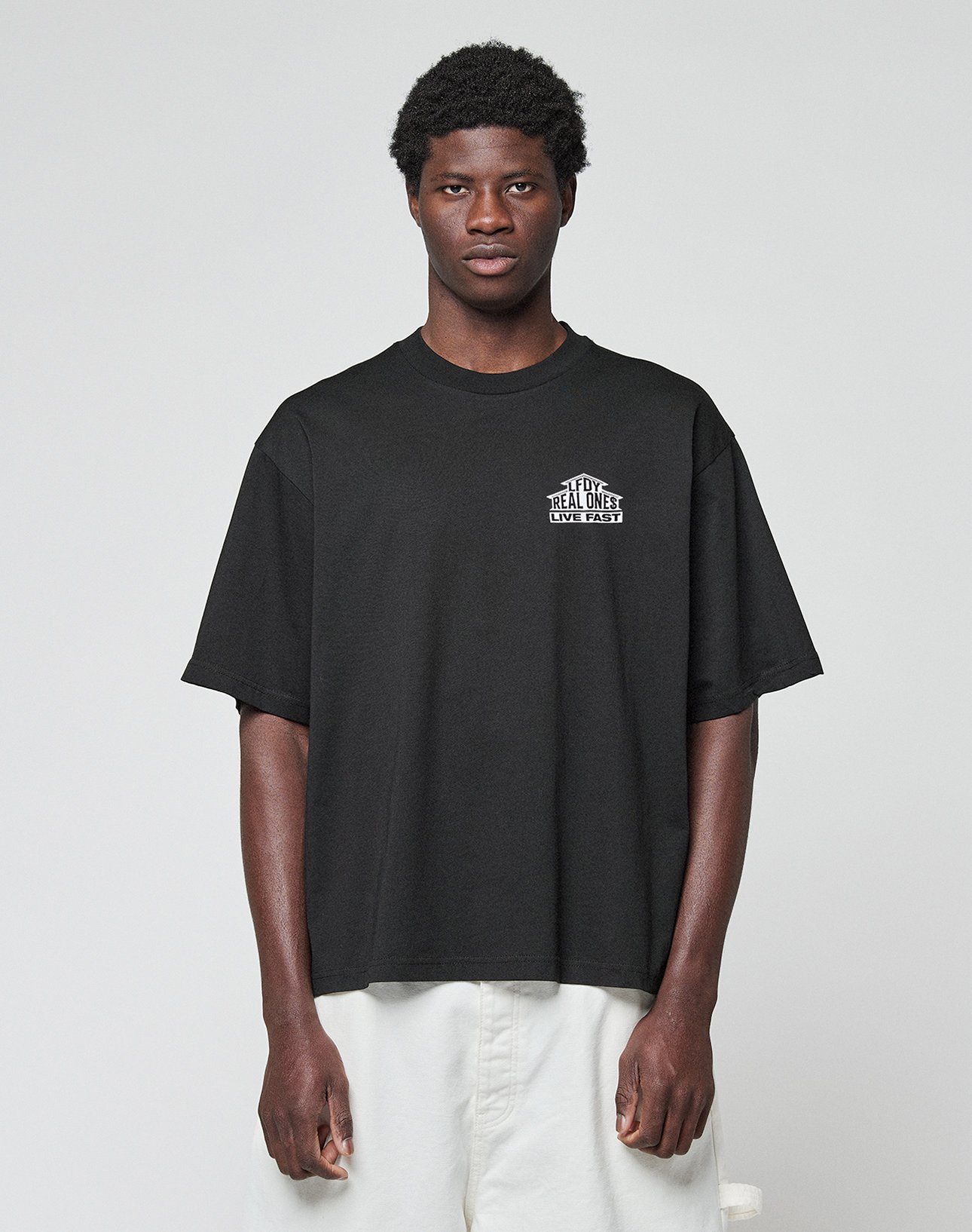 A young man with short curly hair wears the LIVE FAST DIE YOUNG LFDY Real Ones Tee in Oversize Fit with white chest text and white shorts, standing against a plain light gray background.