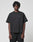 A person with short dreadlocks wears the LIVE FAST DIE YOUNG Basic Trademark Tee in an oversize fit with black pants, both made from 100% Baumwolle, standing against a plain light gray background facing the camera neutrally.