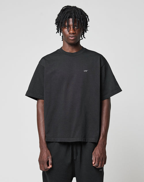 A person with short dreadlocks wears the LIVE FAST DIE YOUNG Basic Trademark Tee in an oversize fit with black pants, both made from 100% Baumwolle, standing against a plain light gray background facing the camera neutrally.