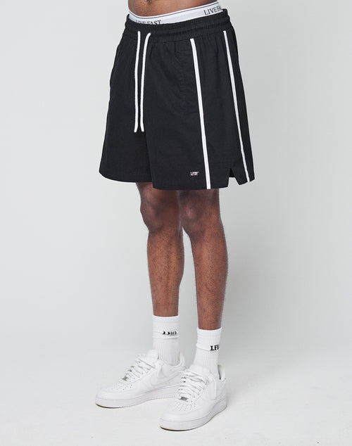 A person wearing LIVE FAST DIE YOUNG Basic Cotton Shorts—black with white vertical stripes, elastic waistband, regular fit—paired with white socks and sneakers, stands against a plain white background.