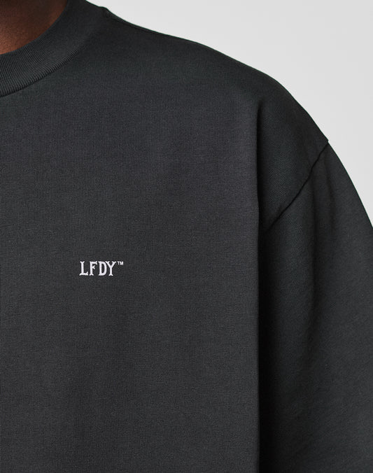 Close-up of a person wearing the LIVE FAST DIE YOUNG Basic Trademark Tee. The black crewneck features white embroidered LFDY™ on the chest. Only part of the shoulder, neck, and upper chest is visible. Made from 100% Baumwolle.