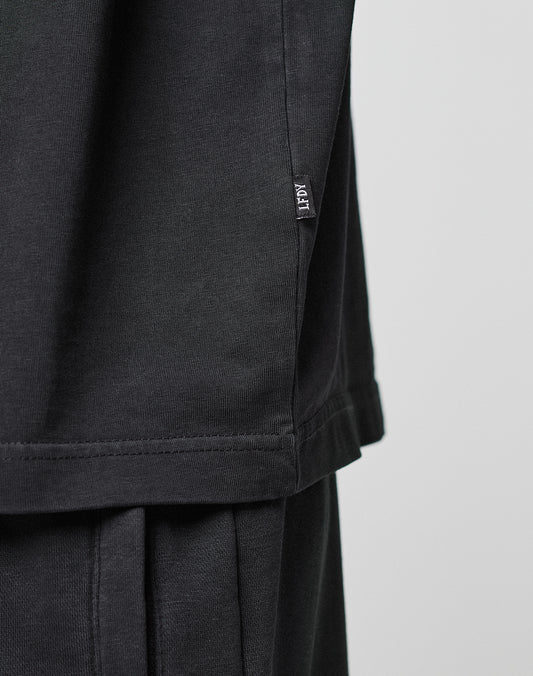 Close-up of a person wearing the LIVE FAST DIE YOUNG Basic Trademark Tee in black with an oversize fit, paired with black pants. A small black ACGT tag is visible at the hem. The background is plain and light-colored.