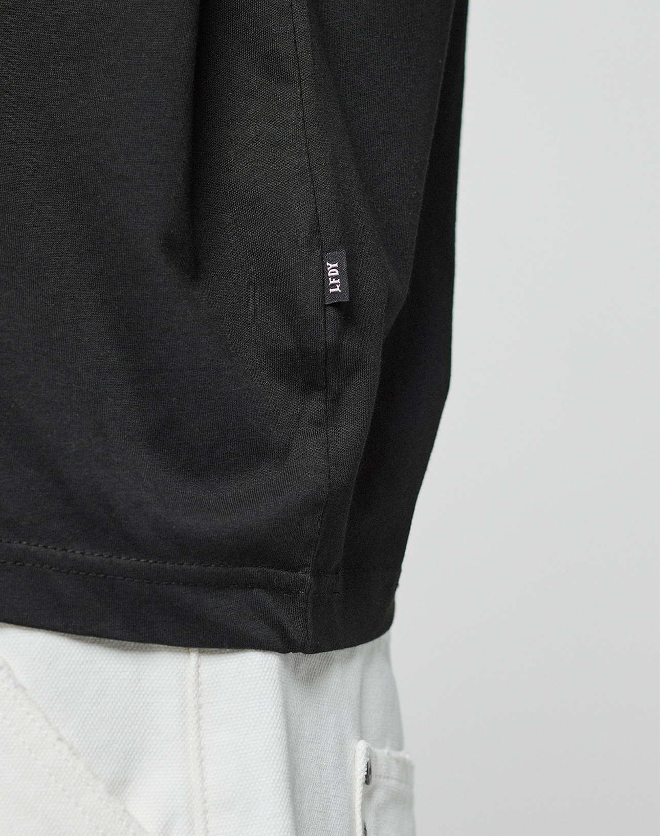 Close-up of a person wearing the LFDY Real Ones Tee by LIVE FAST DIE YOUNG in black, made from 100% Baumwolle, paired with light pants. The lower shirt features a small black tag with white text against a plain background.