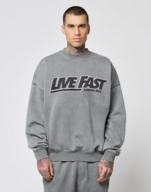 A man with short dark hair and hand tattoos faces forward, wearing the LIVE FAST DIE YOUNG Live Fast Europe Run Sweater in oversize fit with a bold frontprint. The background is plain white. | model
