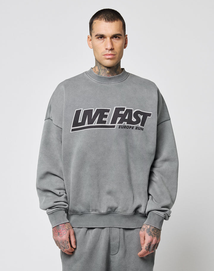 A man with short dark hair and hand tattoos faces forward, wearing the LIVE FAST DIE YOUNG Live Fast Europe Run Sweater in oversize fit with a bold frontprint. The background is plain white. | model