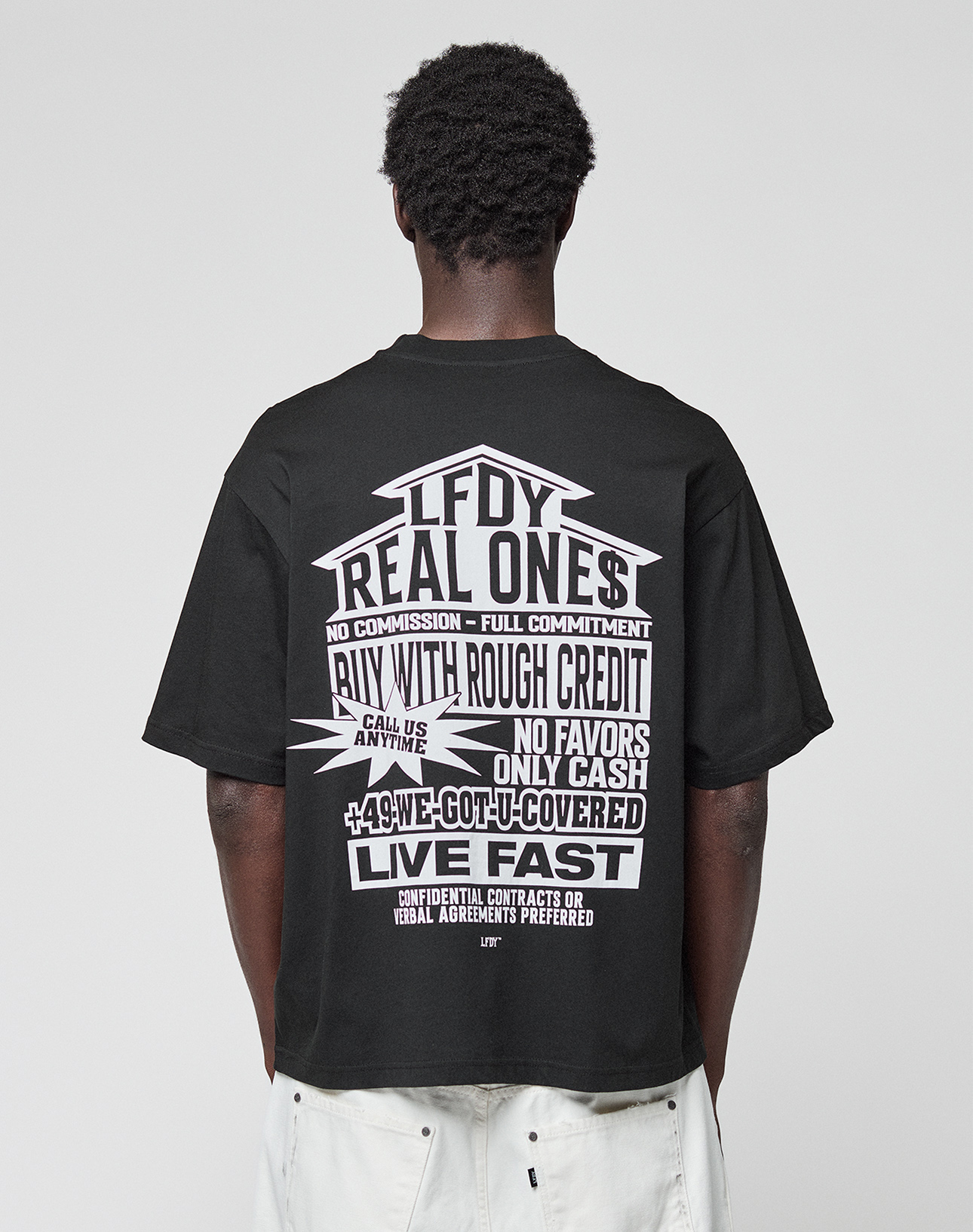 A person with short curly hair, seen from behind, wears the LFDY Real Ones Tee by LIVE FAST DIE YOUNG—a loose black 100% Baumwolle T-shirt with bold white "Real Ones" graphic—paired with loose white shorts.