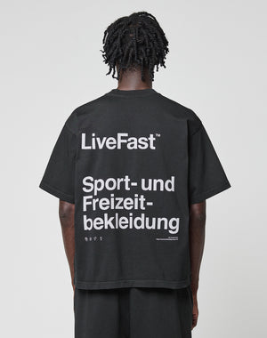A person with short, twisted hair is seen from behind wearing a black LIVE FAST DIE YOUNG Basic Trademark Tee with large white “LiveFast™ Sport- und Freizeitbekleidung” text. The background is plain and light-colored.