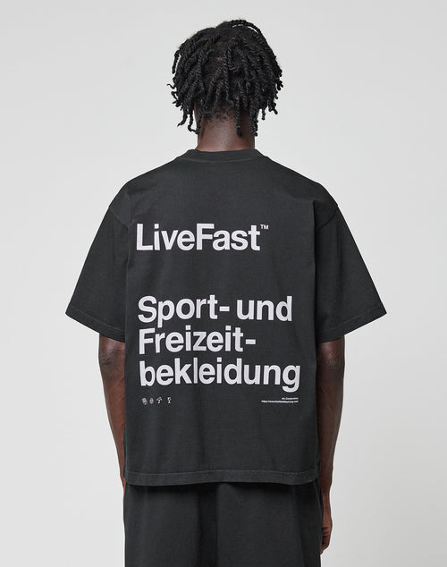 A person with short, twisted hair is seen from behind wearing a black LIVE FAST DIE YOUNG Basic Trademark Tee with large white “LiveFast™ Sport- und Freizeitbekleidung” text. The background is plain and light-colored.
