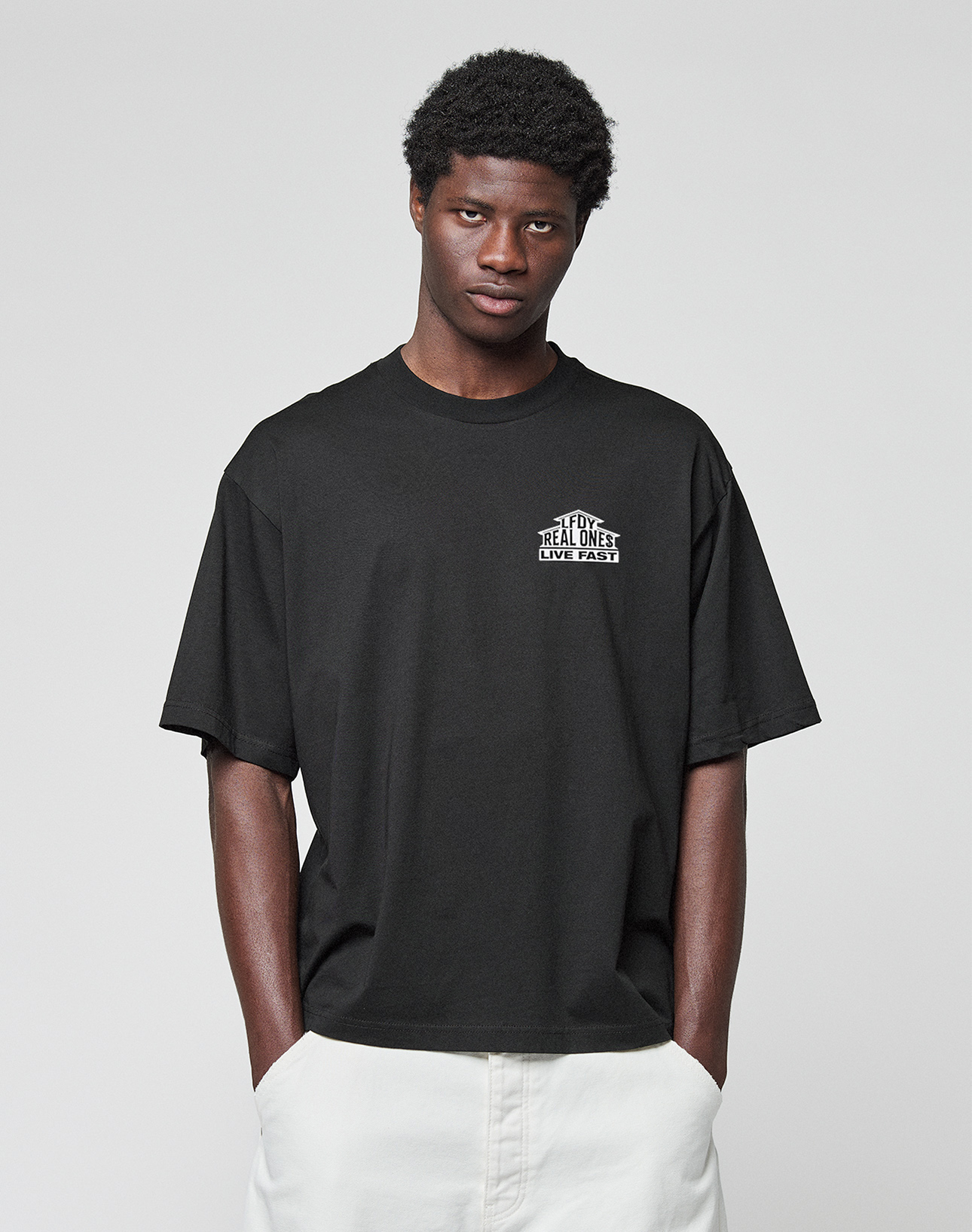 A person wears the LIVE FAST DIE YOUNG LFDY Real Ones Tee, a loose black T-shirt with front and back print, and white pants, standing against a plain light gray background with hands in pockets, facing the camera.