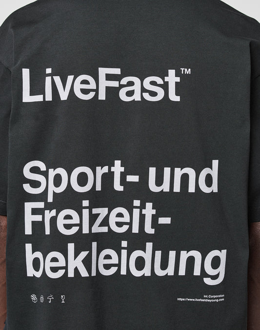 A close-up of the back of the black LIVE FAST DIE YOUNG Basic Trademark Tee shows large white text: LiveFast™ Sport- und Freizeitbekleidung. Small icons and a URL appear at the bottom. 100% Baumwolle, markanter Front- und Backprint.