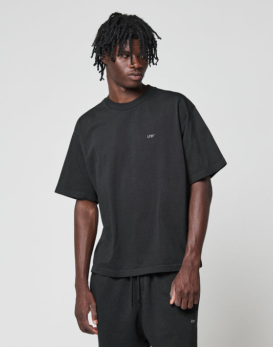 A man with short dreadlocks wears the LIVE FAST DIE YOUNG Basic Trademark Tee, a loose-fitting black T-shirt with front and back print, oversize fit, and 100% cotton, paired with black shorts against a light gray background.