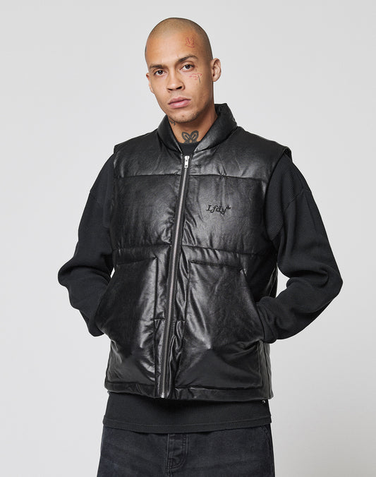A person with a shaved head and face tattoos wears the LIVE FAST DIE YOUNG Padded Vest in black, regular fit, with padded stand-up collar over a black pullover, hands casually in pockets, standing against a plain light gray background.
