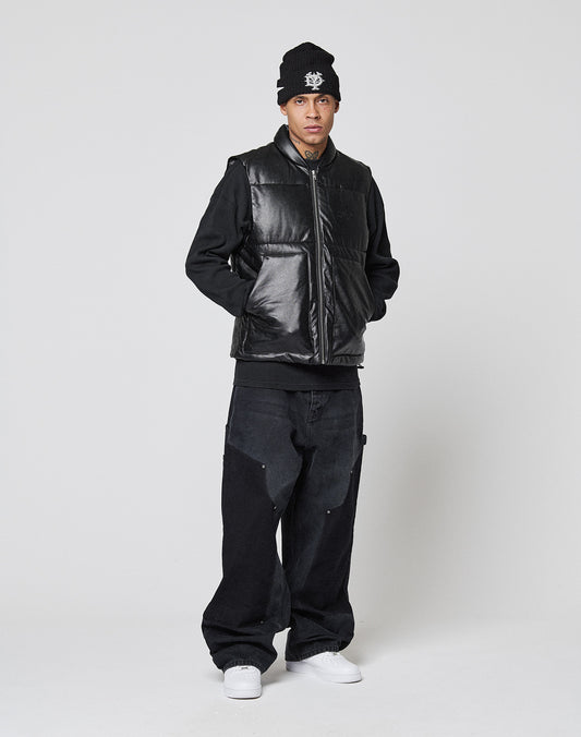 A person stands against a plain background wearing a black beanie, LIVE FAST DIE YOUNG Padded Vest with a stand collar, black long-sleeve shirt, loose regular fit black jeans, and white sneakers, looking at the camera with hands in the vest pockets.